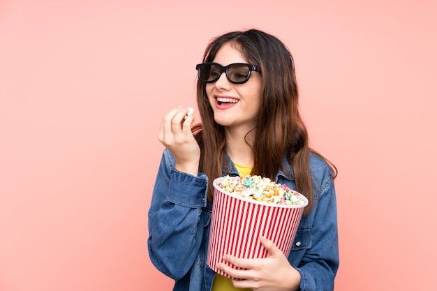 Jonge donkerbruine vrouw over geïsoleerde roze muur met 3d glazen en holding een grote emmer popcorns