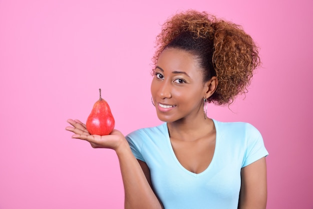 Jonge donkere vrouw houdt een rode peer in de palm van haar hand en glimlacht op een roze achtergrond.