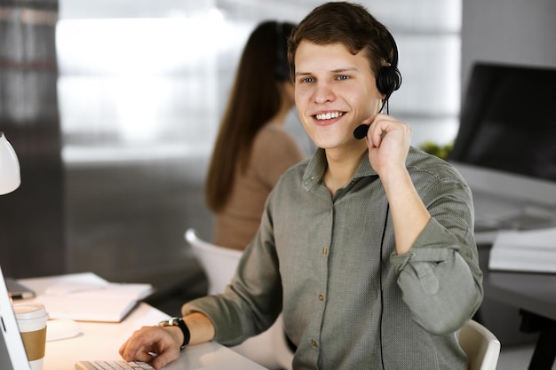 Jonge donkerharige man in een groen shirt en headsets praat met een klant terwijl hij aan het bureau zit en samenwerkt met een vrouwelijke collega in een modern kantoor Callcenter operators op het werk