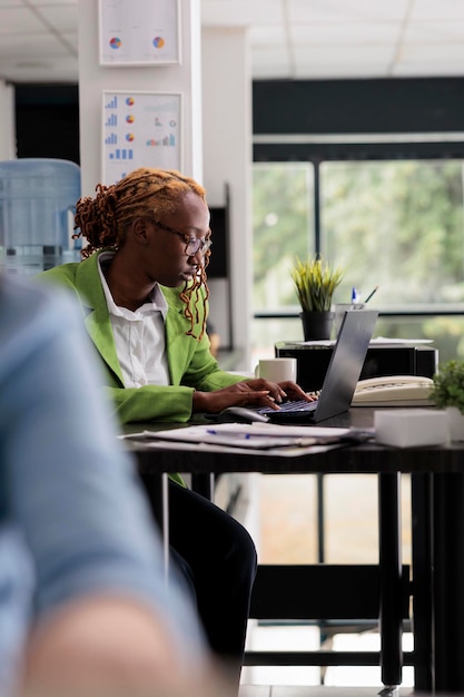 Jonge doordachte werknemer die op laptop werkt in een klein kantoor, professionele bedrijfsmanager die aan het bureau van de werkplek zit, zijaanzicht. Gerichte Afro-Amerikaanse werknemer, werkdruk