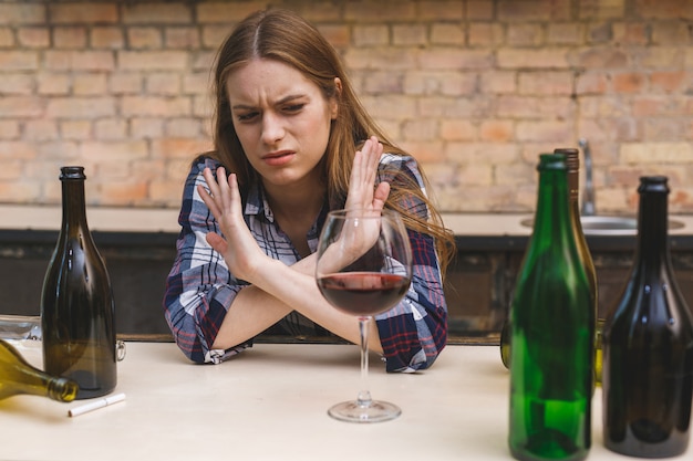 Jonge droevige en verspilde alcoholische vrouw die aan keukenbank zit die rode wijn drinkt, volledig dronken die depressief eenzaam kijkt en kater lijdt in alcoholisme en alcoholmisbruik.