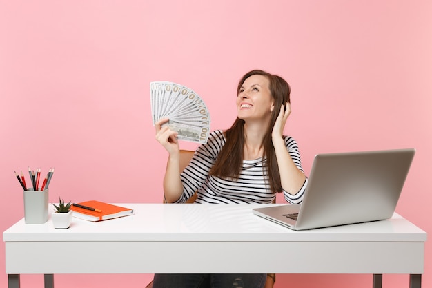 Jonge dromerige glimlachende vrouw die denkt dat ze een bundel met veel dollars vasthoudt, contant geld werkt aan een wit bureau met een pc-laptop