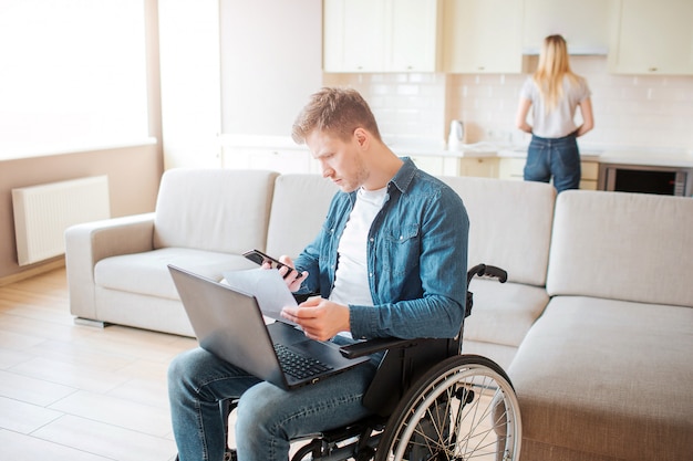 Jonge drukke man met een handicap zittend op rolstoel. Houd laptop op knieën. Jonge vrouw achter staan en koken. Daglicht in de kamer.