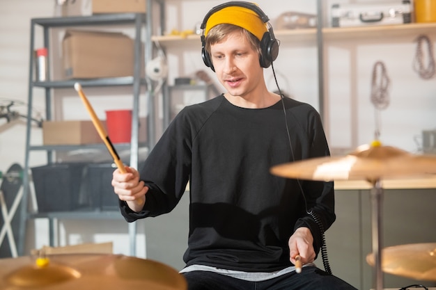 Jonge drummer met koptelefoon die drumstokken boven cimbalen houdt terwijl hij een van hen gaat verslaan tijdens individuele repetitie