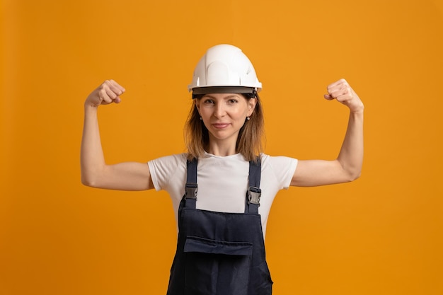 Foto jonge dunne vrouwenbouwer in een bouwvakker op een oranje achtergrond toont biceps