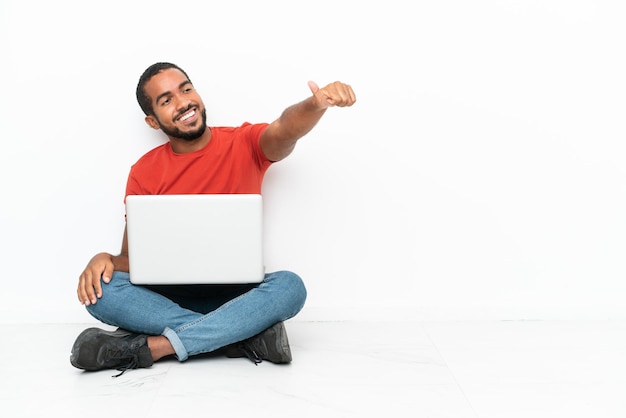 Jonge Ecuadoraanse man met een laptop zittend op de vloer geïsoleerd op een witte achtergrond met een duim omhoog gebaar