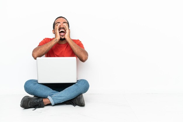 Jonge Ecuadoraanse man met een laptop zittend op de vloer geïsoleerd op een witte achtergrond schreeuwen en iets aankondigen