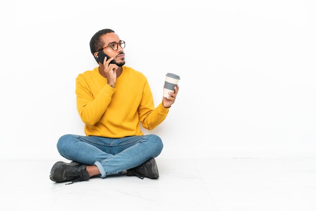 Jonge Ecuadoraanse man zittend op de vloer geïsoleerd op een witte muur met koffie om mee te nemen en een mobiel