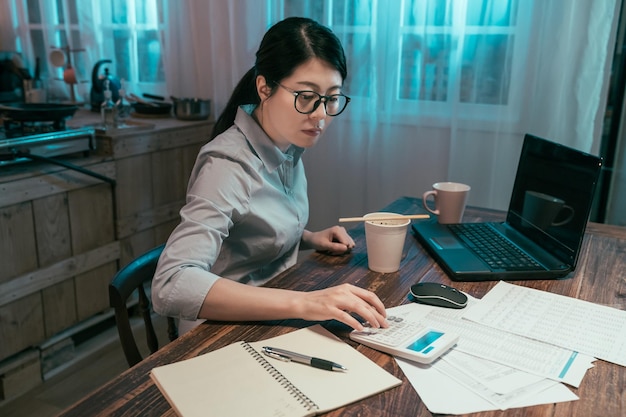 jonge elegante Aziatische Koreaanse zakenvrouw die de financiering van het bedrijf telt op rekenmachine met laptopcomputer en documentpapieren op tafel in de thuiskeuken. kantoordame geniet van instant noedels tijdens het werken