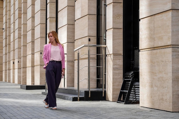 Jonge elegante mooie blonde duizendjarige met lang haar in roze kleding loopt in de buurt van een modern gebouw Volledige lengte wissen Selectieve focus