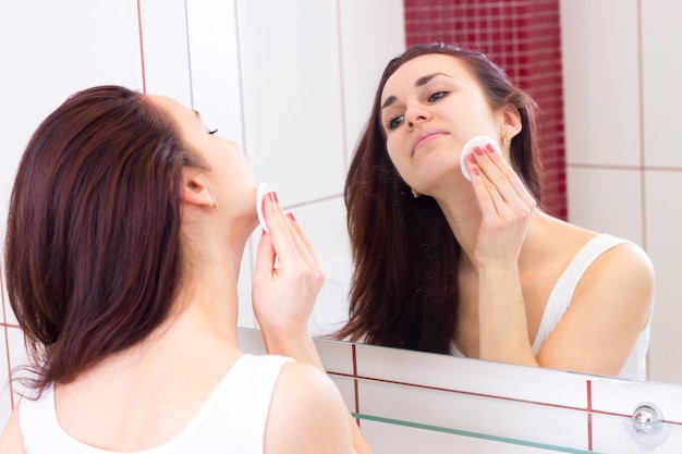 Jonge elegante vrouw die make-up verwijdert met een wit wattenschijfje voor de spiegel in haar badkamer