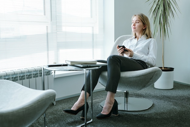 Jonge elegante zakenvrouw met mobiele telefoon die door het raam kijkt terwijl ze in een zachte comfortabele fauteuil aan een kleine tafel zit