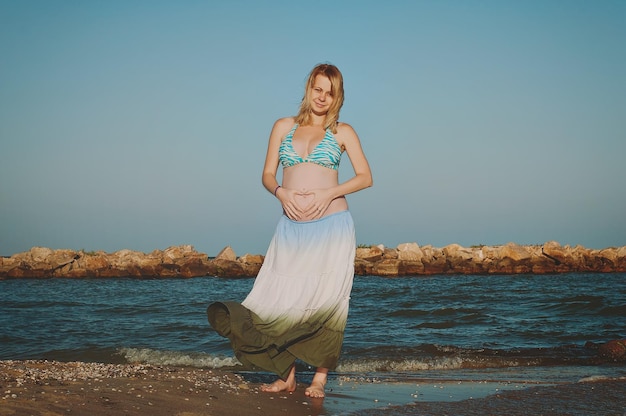 jonge elegante zwangere blonde vrouw in een blauwe hoed loopt op het strand bij zonsondergang. Zee van Azov. Moederschap