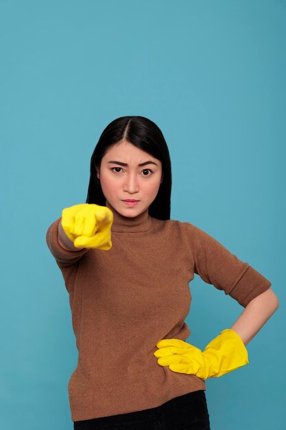 Jonge emotionele en ongelukkige Aziatische huishoudster van klusjes wijzende vinger naar voren geïsoleerd op een blauwe achtergrond, schoonmaak thuisconcept, overwerk uitgeputte vrouw met negatieve gemoedstoestand