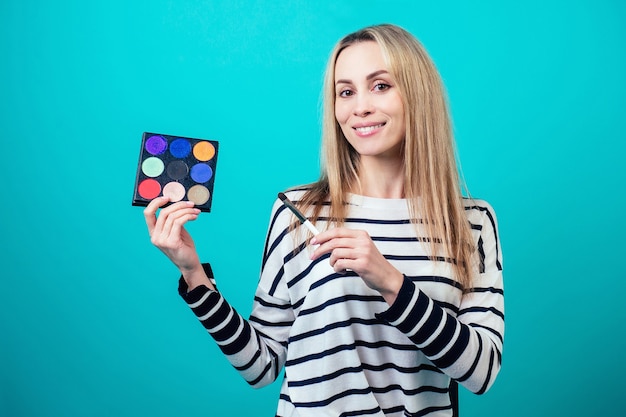 Jonge en aantrekkelijke blonde vrouw visagiste visagist houdt een borstel make-up en een palet van oogschaduw op blauwe achtergrond in een studio. concept van huidverzorging en schoonheidscosmetica voor gezicht.