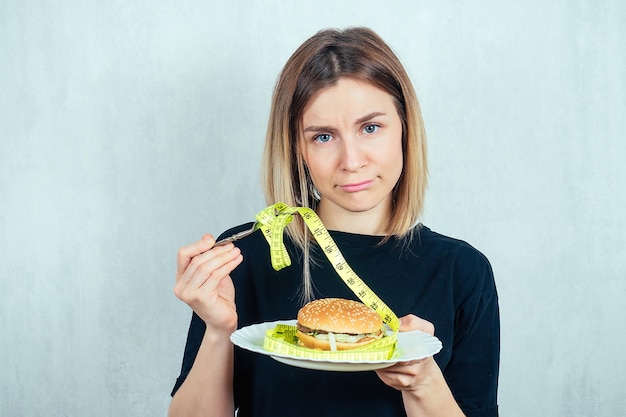 Jonge en aantrekkelijke trieste blonde vrouw in zwarte T-shirt en meetlint houdt een calorieburger op een bordconcept van afwijzing van schadelijk fastfood en dieet