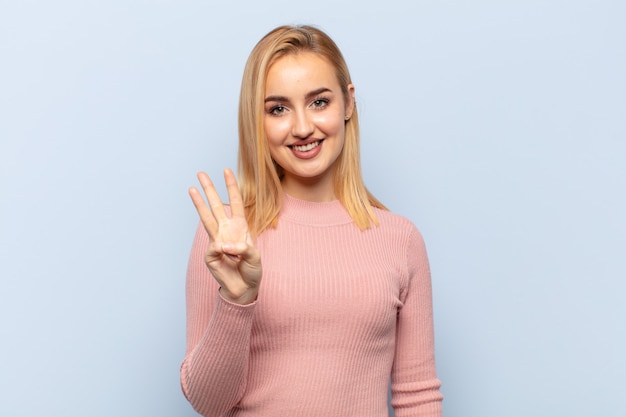 Jonge en blonde vrouw die vriendelijk glimlacht kijkt, nummer drie of derde met vooruit hand toont, aftellend