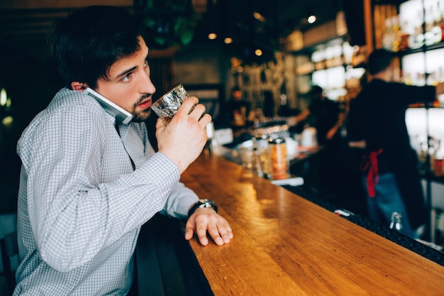 Jonge en donkerharige man zit bij de kelner in de club en drinkt wat alcohol. Ook praat hij aan de telefoon en probeert hij tegelijkertijd te drinken. Barman staat ver van hem af.