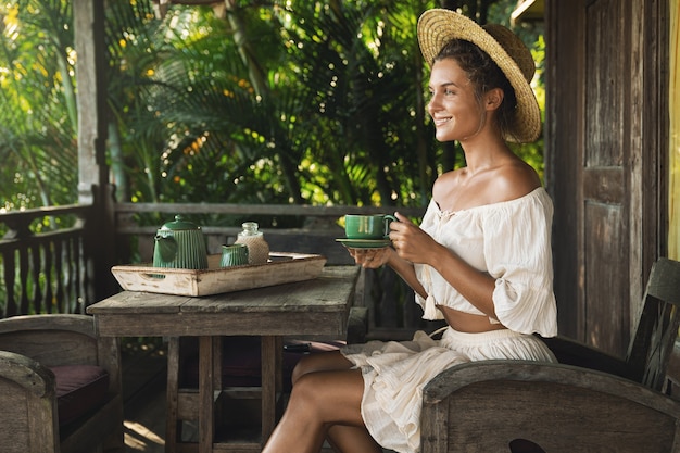 Jonge en gelukkige vrouw zittend op het zomerterras en koffie drinken