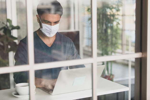 Jonge en knappe blanke zakenman met een beschermend masker voor medische hygiëne die in huis zit, koffie drinkt en een laptop-notebookcomputer gebruikt. Idee om thuis te werken in de coronacrisis.