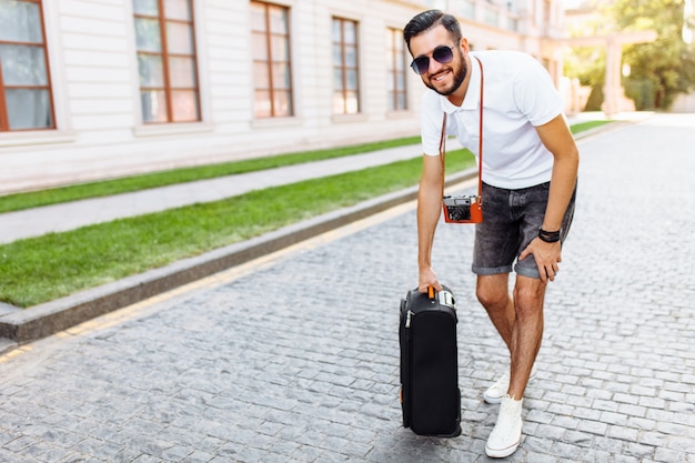 Jonge en knappe man toerist met een baard, wandelen door de stad met een koffer en een camera op zijn nek