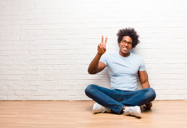 Jonge en mens die gelukkig, onbezorgd en positief glimlacht kijkt, overwinning of vrede gesturing met één hand thuis zittend op de vloer