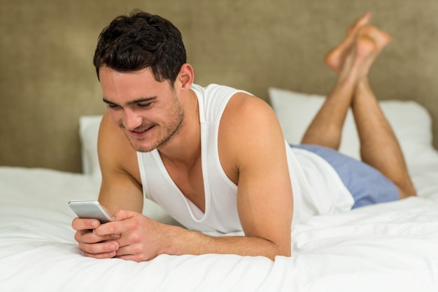 Jonge en mens die terwijl het liggen op bed met zijn telefoon texting glimlachen