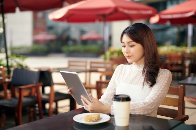 Jonge en mooie Aziatische vrouw die haar tablet-pc gebruikt terwijl ze in de buitencafé in de stedelijke stad zit