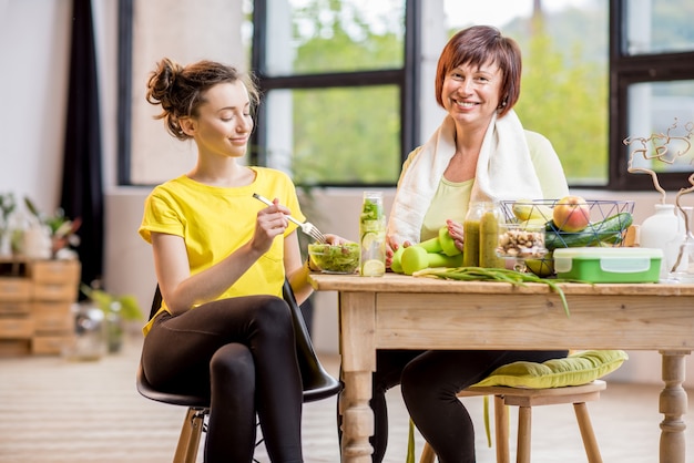 Jonge en oudere vrouwen oefenen met halters en eten gezond voedsel binnenshuis op de achtergrond van het raam