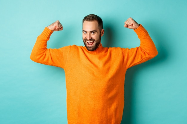 Jonge en sterke man kijkt zelfverzekerd, buigt biceps en pronkt met spierkracht, glimlachend zelfverzekerd, staande over lichtblauwe achtergrond.