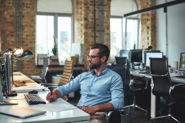 Jonge en succesvolle bebaarde man in bril en formele kleding kijken naar computermonitor