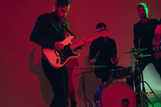 Jonge en vrolijke kaukasische muzikanten die optreden op rode studioachtergrond in neonlicht. Concept van muziek, hobby, festival. Kleurrijk portret van moderne kunstenaar. Betrokken en geïnspireerd. Kunst, coverband.