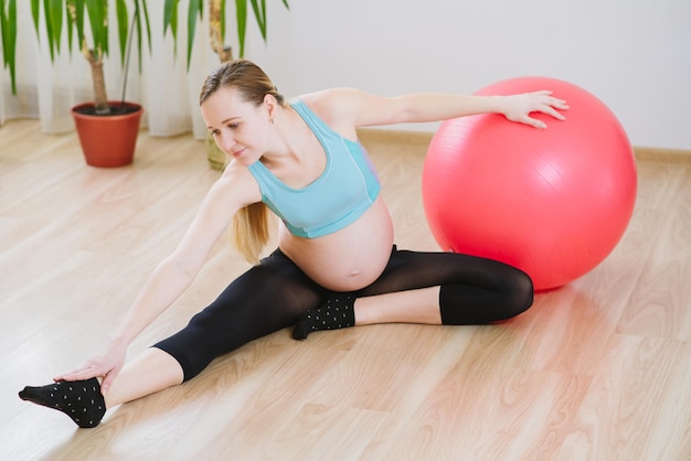 Jonge en zwangere vrouw in sportpak die thuis oefeningen doet met een grote bal