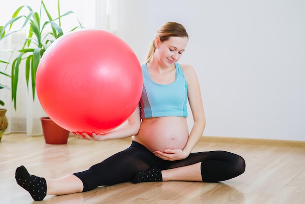 Jonge en zwangere vrouw in sportpak die thuis oefeningen doet met een grote bal