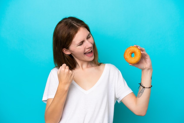 Jonge engelse vrouw die een doughnut over geïsoleerde blauwe achtergrond houdt die een overwinning viert