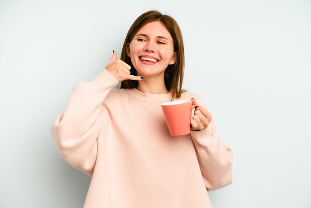 Jonge Engelse vrouw die een mok houdt die op blauwe achtergrond wordt geïsoleerd die een mobiel telefoongesprekgebaar met vingers toont