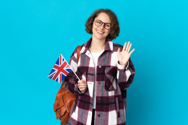 Jonge Engelse vrouw die een vlag van het Verenigd Koninkrijk houdt die op blauwe muur wordt geïsoleerd die met hand met gelukkige uitdrukking groet