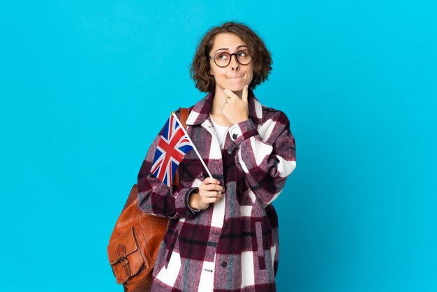 Jonge Engelse vrouw die een vlag van het Verenigd Koninkrijk houdt die op blauwe muur wordt geïsoleerd die twijfels heeft en denkt