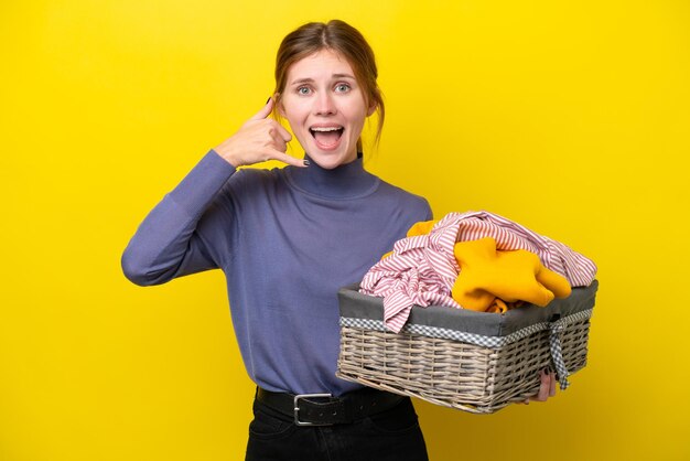 Jonge Engelse vrouw die een wasmand houdt die op gele achtergrond wordt geïsoleerd en telefoongebaar maakt Bel me terug-teken