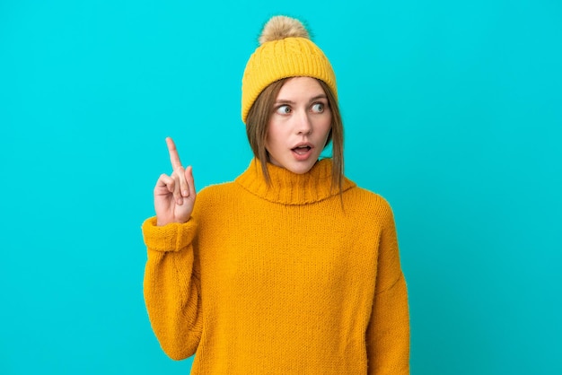 Jonge Engelse vrouw die een winterjas draagt die op een blauwe achtergrond wordt geïsoleerd en een idee denkt dat de vinger omhoog wijst