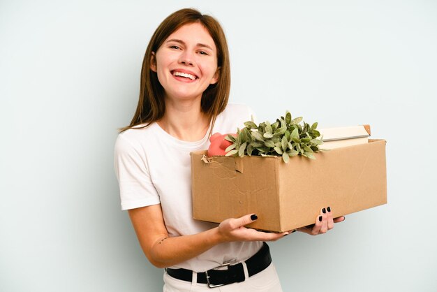 Jonge Engelse vrouw die een zet doet terwijl ze een doos vol dingen oppakt die geïsoleerd zijn op een blauwe achtergrond, lachen en plezier hebben
