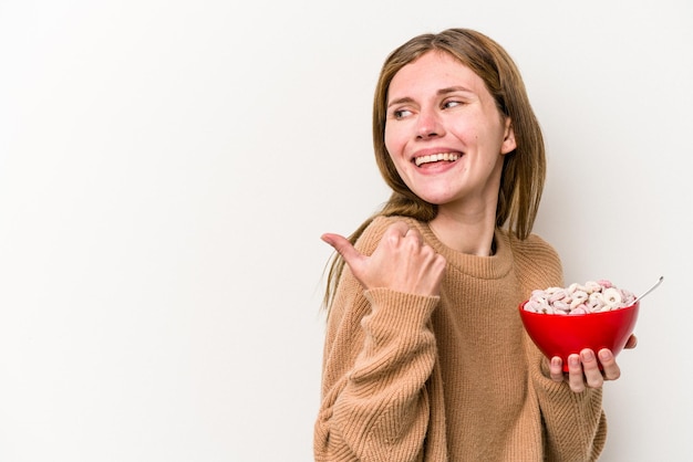 Jonge Engelse vrouw die granen eet die op een witte achtergrond wordt geïsoleerd, wijst met duimvinger weg, lachend en zorgeloos.