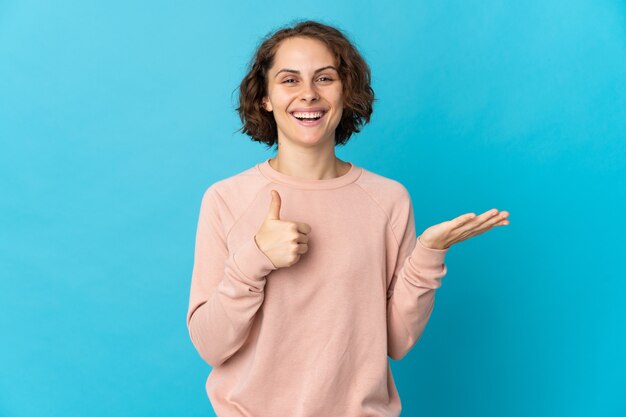 Jonge Engelse vrouw die op blauwe muur wordt geïsoleerd die copyspace denkbeeldig op de palm houdt om een advertentie en met omhoog duimen in te voegen