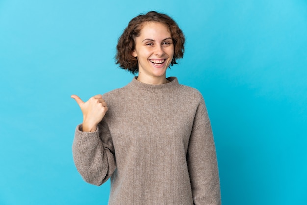 Jonge Engelse vrouw die op blauwe muur wordt geïsoleerd die naar de kant wijst om een product te presenteren