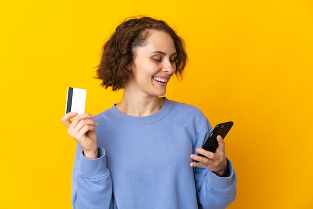 Jonge Engelse vrouw die op gele muur wordt geïsoleerd die met mobiel met een creditcard koopt