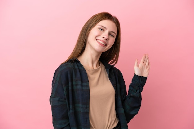 Jonge Engelse vrouw die op roze achtergrond wordt geïsoleerd die met hand met gelukkige uitdrukking salueert