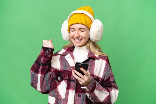 Jonge Engelse vrouw die wintermoffen draagt over geïsoleerde achtergrond met telefoon in overwinningspositie