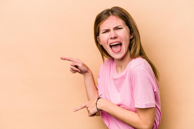 Jonge Engelse vrouw geïsoleerd op beige achtergrond wijzend met wijsvingers naar een kopieerruimte die opwinding en verlangen uitdrukt