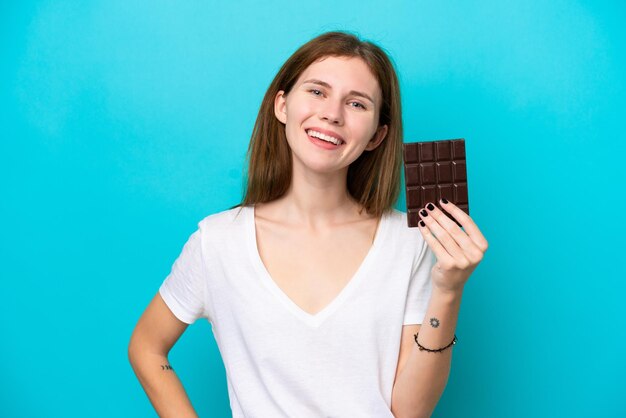 Jonge Engelse vrouw geïsoleerd op blauwe achtergrond die een chocoladetablet neemt en happy