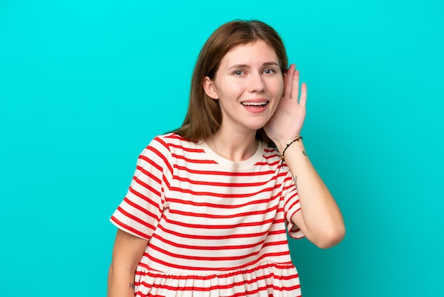 Jonge Engelse vrouw geïsoleerd op een blauwe achtergrond die naar iets luistert door hand op het oor te leggen